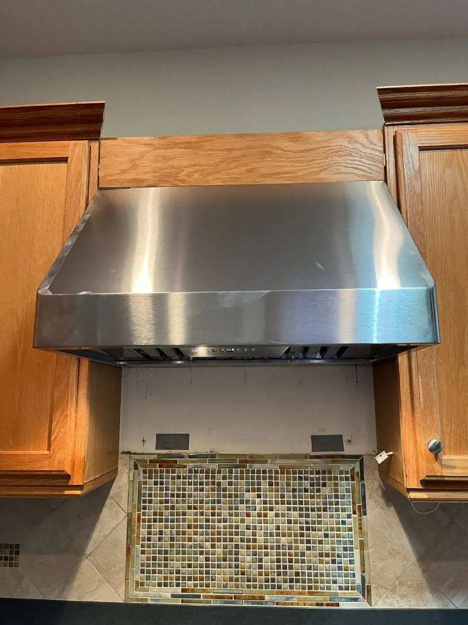 Stainless steel range hood mounted under oak cabinets above a mosaic tile backsplash