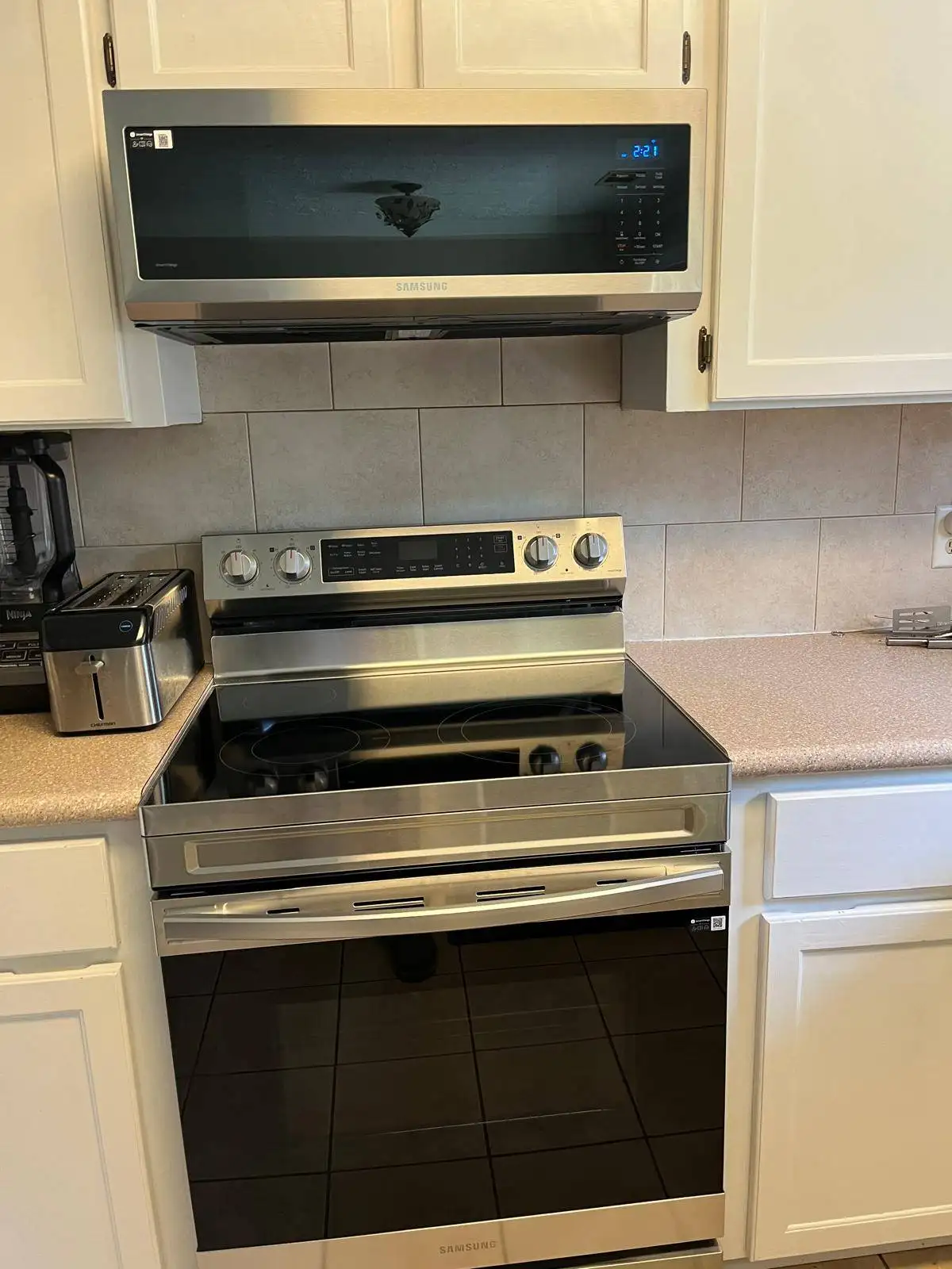 Stainless steel Samsung over-the-range microwave and electric range with a glass cooktop in a kitchen with white cabinets.