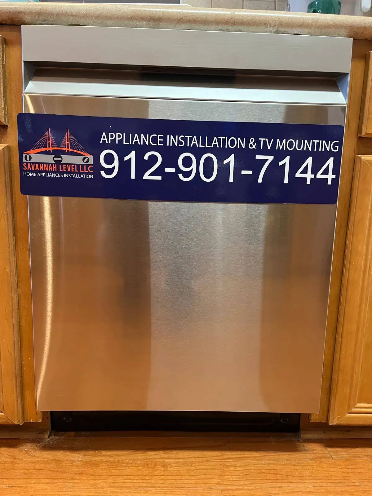 Stainless steel built-in dishwasher with a Savannah Level LLC appliance installation and TV mounting magnet, between wooden kitchen cabinets.