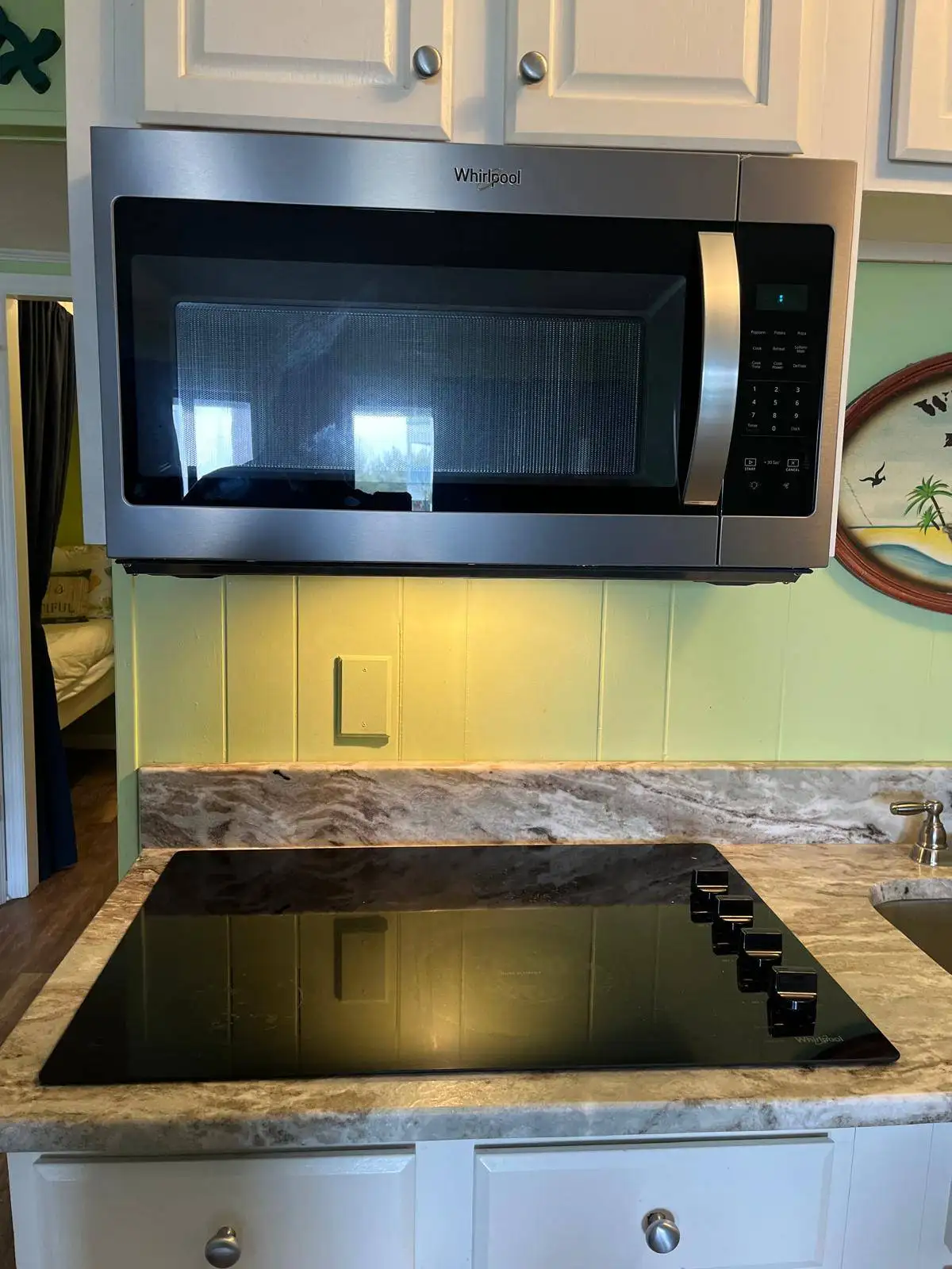 Stainless steel Whirlpool over-the-range microwave above a black glass electric cooktop in a kitchen with white cabinets and a marble-look countertop.